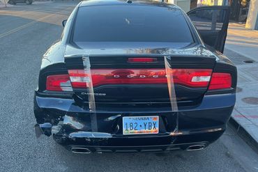Damaged black Dodge Charger with taped rear bumper on a street.