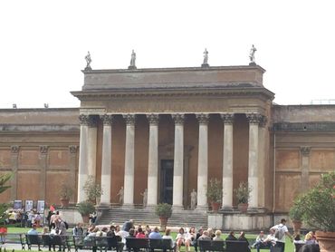 Ancient building with tall columns and statues, people sitting on benches in front.
