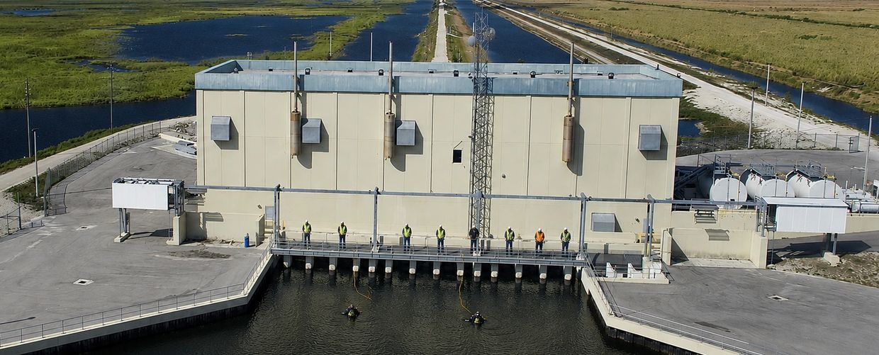 Workers stand on a water facility with two divers in the water and marshland in the background.