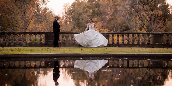 Bride and groom by pond with autumn trees reflecting in water.