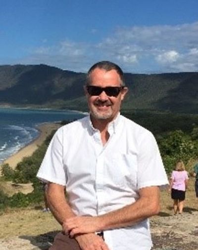 Kim Agnew white man with short grey hair, sunglasses and grey facial hair in front of a beach and mo