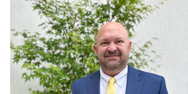 Smiling man in blue suit and yellow tie stands outdoors in front of green foliage.
