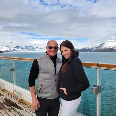 A couple smiles on a cruise deck with snowy mountains and calm water in the background.