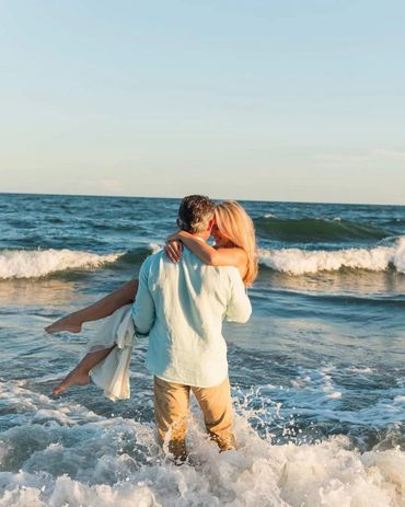 A man carrying a woman in the ocean waves at sunset.