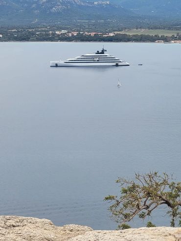 A large white ultra luxury cruise ship and a small sailboat on calm water with a distant shoreline and mountains off the coast of Corsica, FR. Concierge created!
