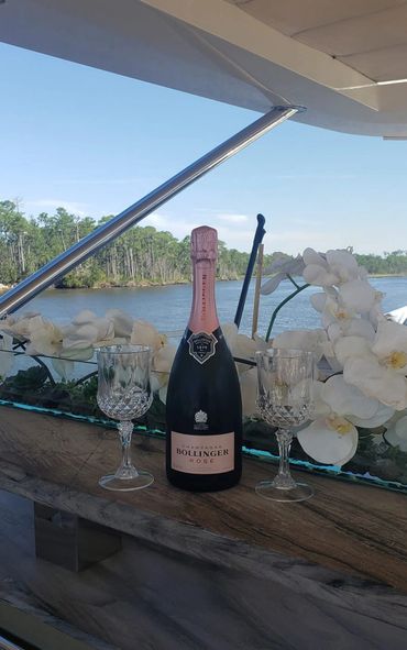 A bottle of Bollinger Rosé champagne with two crystal glasses and white orchids by the water.