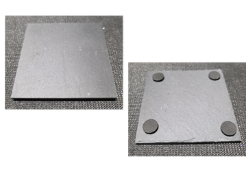 Two black square coasters, one with felt pads on corners, on a black textured surface.