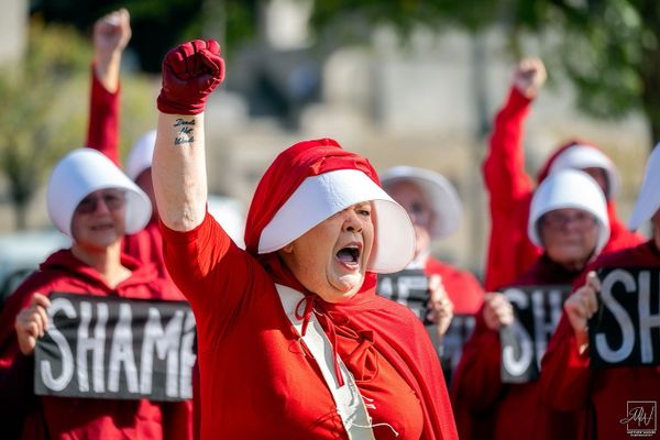 Protesters in red cloaks and white bonnets raise fists, holding 'SHAME' signs.