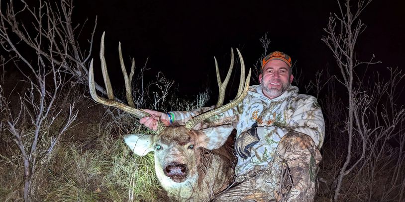 Hunter posing with a large buck deer at night in camo gear.