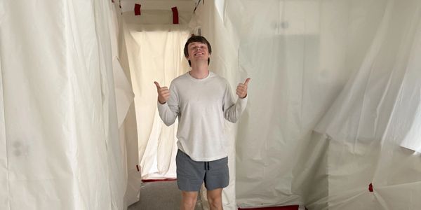 A young man in casual clothes giving thumbs up inside a room covered in white plastic sheets.
