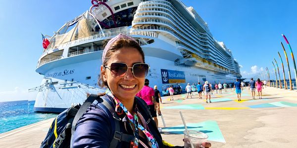 Woman taking a selfie in front of a large cruise ship on a sunny day.