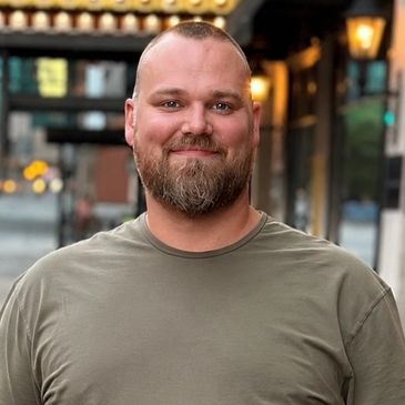 Smiling bearded man in a casual t-shirt outdoors in an urban setting.