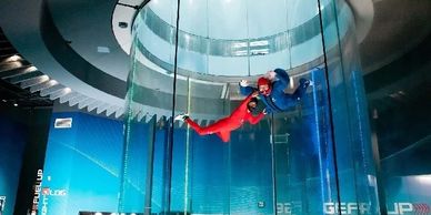 Two people in flight suits indoor skydiving inside a vertical wind tunnel.