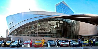 NASCAR drivers with race cars in front of the NASCAR Hall of Fame building.