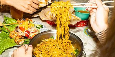 Person eating noodles with chopsticks and another holding flatbread over a table with grilled shrimp and drinks.