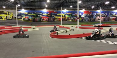 Indoor go-kart track with racers and NASCAR mural.
