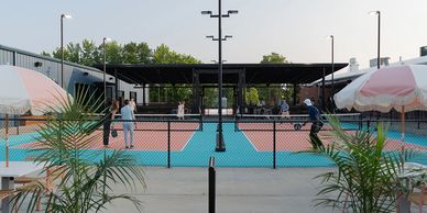 People playing pickleball on outdoor courts with umbrellas and plants nearby.