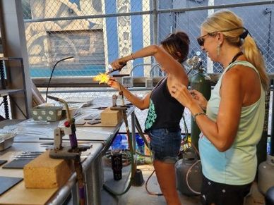 Two women working with glassblowing equipment in a workshop.