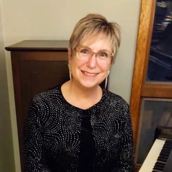 Smiling woman in glasses and sparkly black top sitting by a piano.