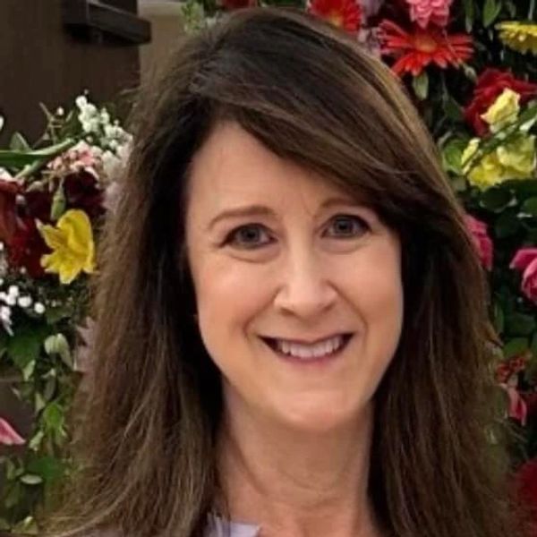 A smiling woman with brown hair stands in front of a colorful floral background.