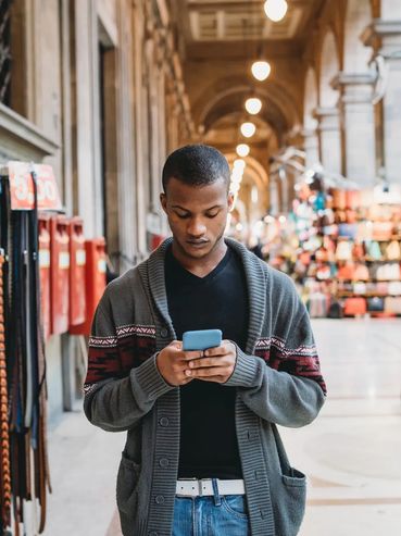 A man using his phone in a bustling market corridor with bags and belts on display.