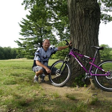 Man in cycling gear with purple mountain bike by a tree outdoors.