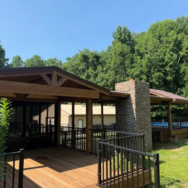 Wooden pavilion with stone fireplace surrounded by greenery under clear sky.