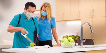 A man with arm sling cuts an apple, assisted by a woman in masks in a modern kitchen.