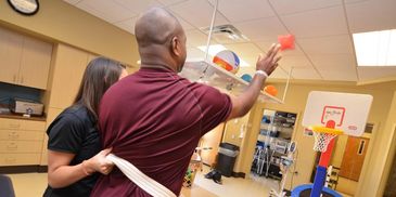 A man playing indoor basketball therapy with assistance from a therapist.