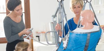Three women operating a patient lift with a mannequin in a wheelchair for training.