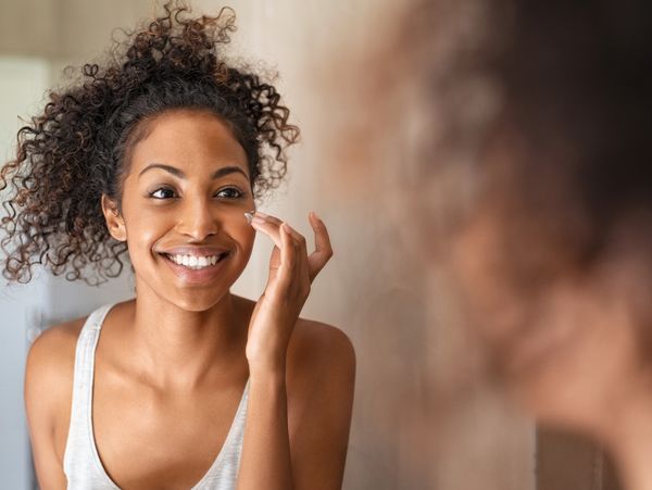 Young black woman applying moisturizer to cheek while standing in front of mirror in bathroom