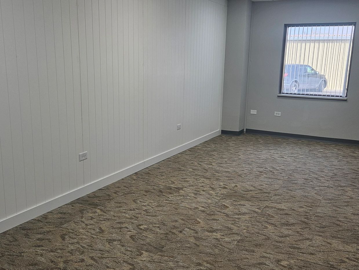 Empty room with carpet, white paneled wall, and a window showing a parked vehicle outside.