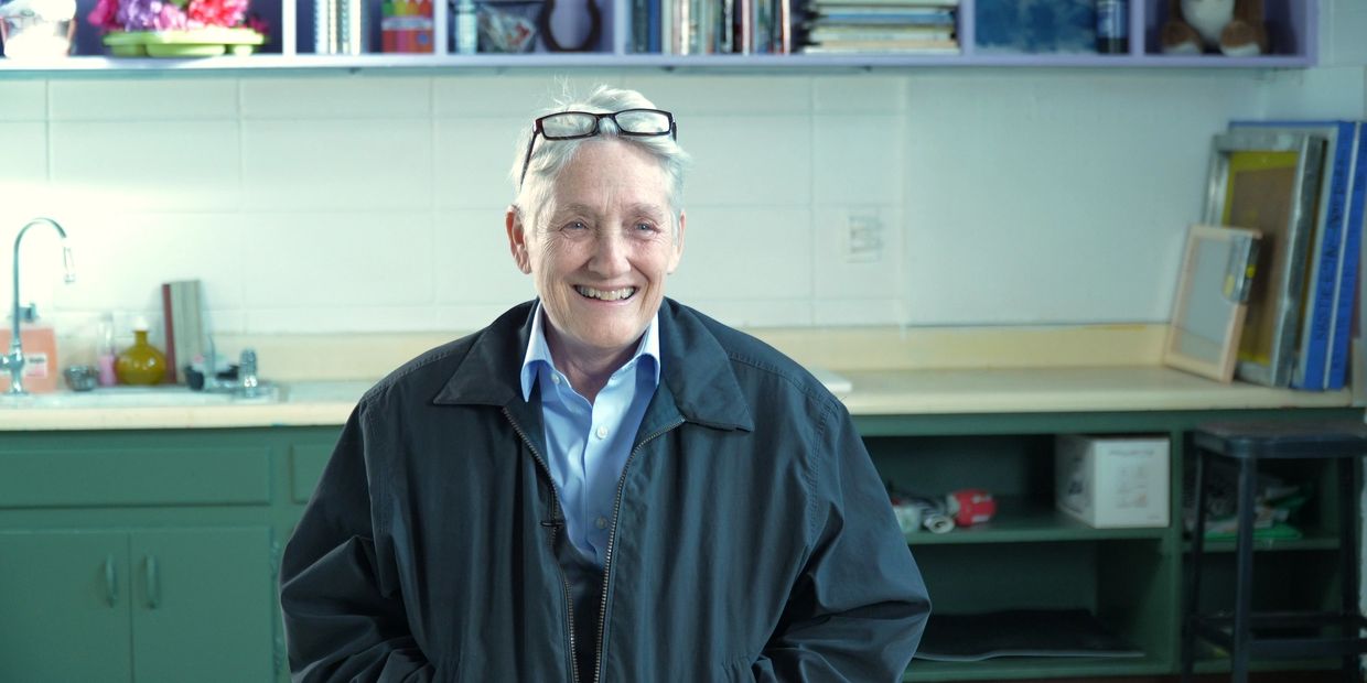Smiling elderly person with glasses on head in a cozy kitchen setting.