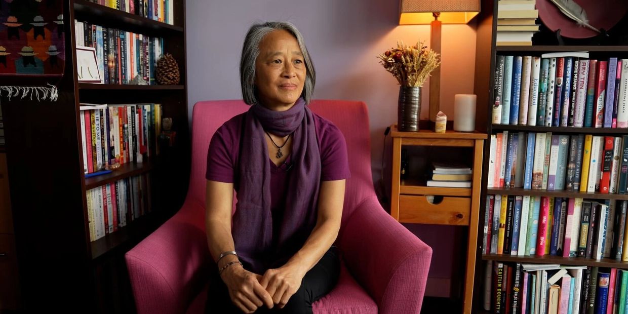 Woman in purple scarf sitting in a pink chair, surrounded by bookshelves and warm lighting.