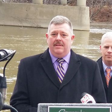 Man in a black coat speaking at an outdoor press conference near a river.