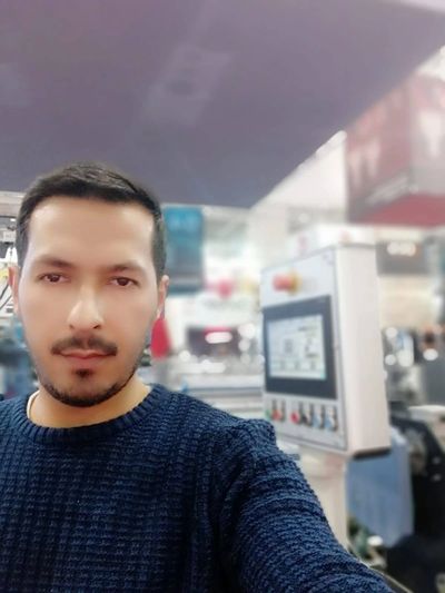 Man in a blue sweater taking a selfie indoors with machinery in the background.