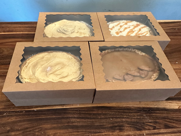 Four assorted pies in brown cardboard boxes with clear windows on a wooden table.