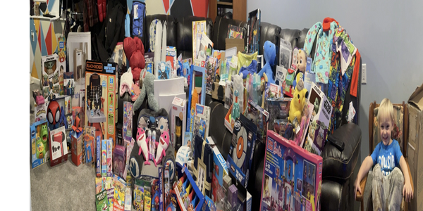 A child sits smiling next to a large collection of toys and gifts in a colorful room.