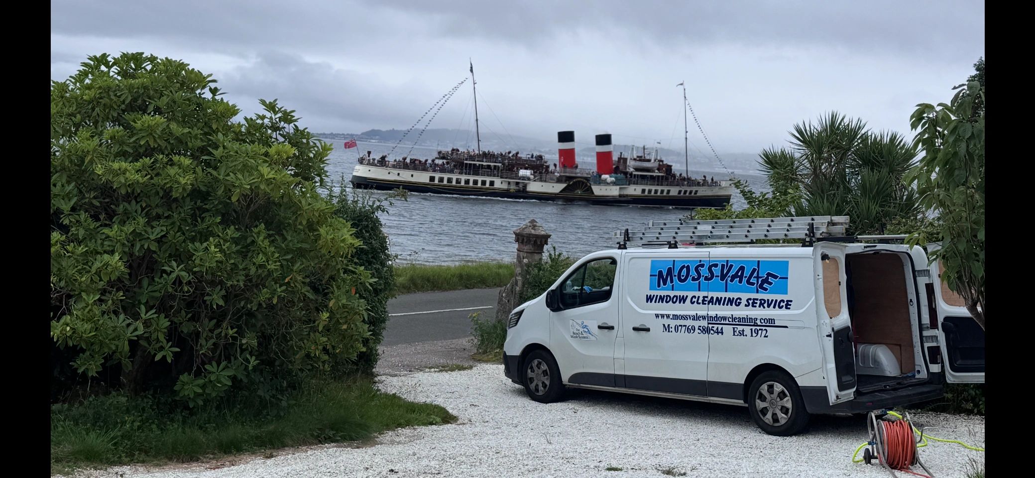Window cleaning van parked near water with a vintage steamboat in the background.