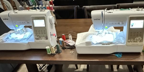 Two Brother sewing machines stitching fabric on a wooden table with colorful thread spools.
