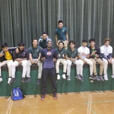 Group of boys posing indoors on a green bench with a coach or teacher in front.