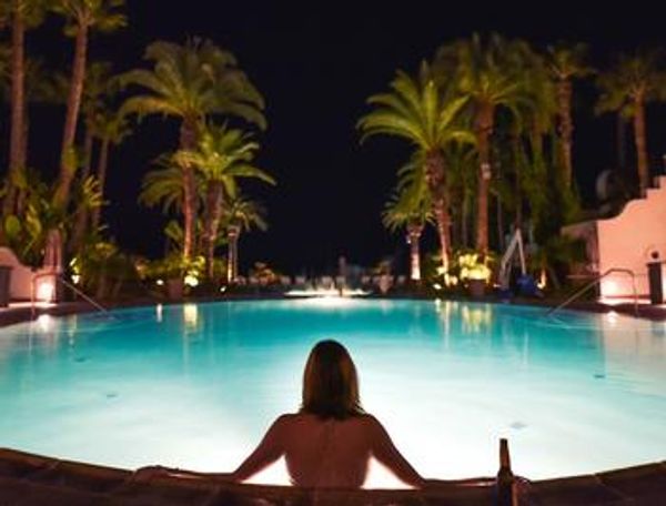 A nighttime view of a person's back as they sit in a pool, surrounded by palm trees.