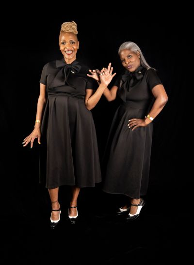 Two women in black dresses holding hands and smiling against a black background.