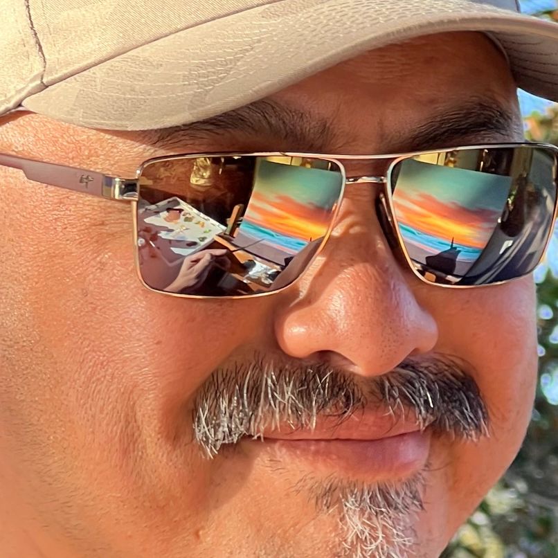 Close-up of a man wearing sunglasses reflecting a vibrant sunset painting.