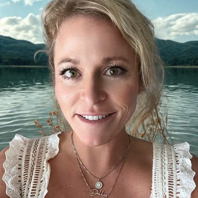 Smiling woman with layered necklaces by a serene lake and mountains.