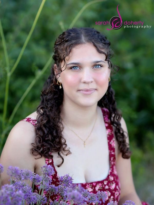 Young woman with curly hair smiles gently among purple flowers in a natural outdoor setting.