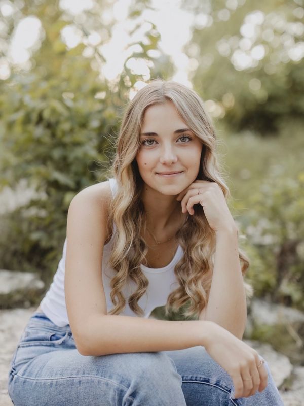 Young woman with long curly hair sitting outdoors, smiling gently.