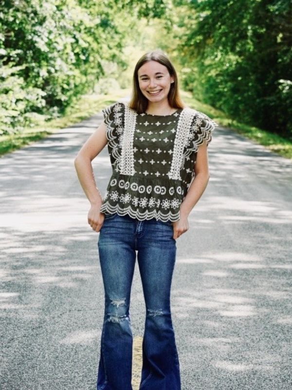 A smiling young woman stands confidently on a sunlit road surrounded by greenery.