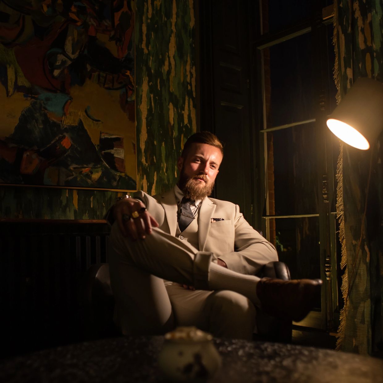 Man in a beige suit sitting confidently in a dimly lit room.