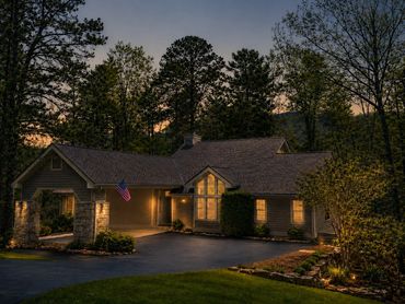 Twilight exterior of Woodsong Lake Toxaway Retreat with warm evening light, wooded privacy, covered arrival, and seasonal mountain-stay appeal.